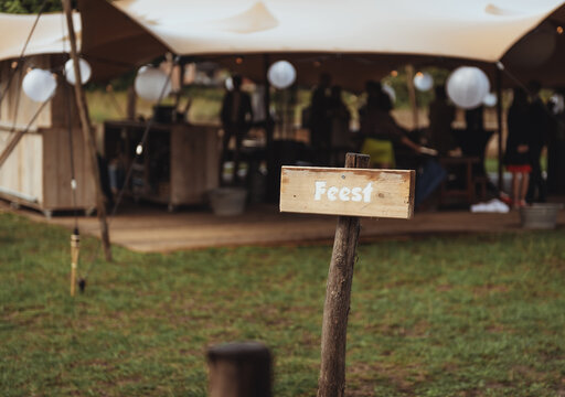 Wooden sign with in white letters in Dutch written "Feest" outdoor in a field in front of a party tent during a festival wedding in summer in The Netherlands with blurry background and space for text.