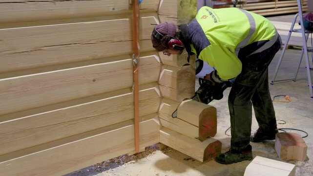 Worker in high-visibility gear uses a chainsaw to cut a corner joint on a log house structure in a woodworking workshop. Indoor setting with wood shavings visible