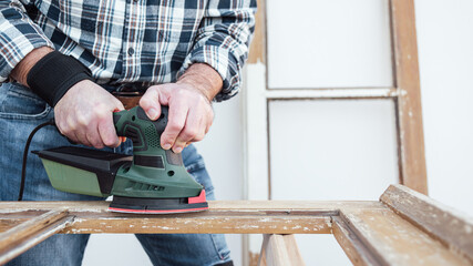 Craftsman. Adult carpenter using an electric sander to smooth an old wooden window. Construction industry, carpentry, housework do it yourself. Restoration.