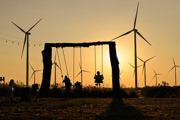 silhouette  of several children playing on swings, with wind turbines in the background.
