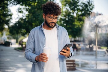Cup of drink and smartphone are in hands. Handsome man is outdoors in the city