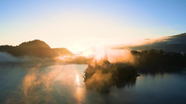 Mari&auml; Himmelfahrt Kirche auf der Insel im See Bled bei Sonnenaufgang, Kamera bewegt sich auf gleicher H&ouml;he durch dichten goldenen Nebel um die Kirche, Geb&auml;ude teilweise im Dunst im warmen Morgenlich