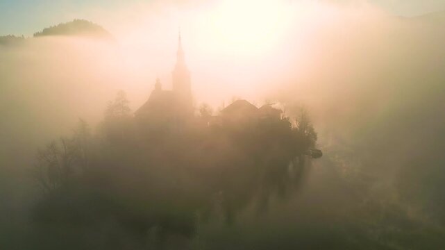 Mari&auml; Himmelfahrt Kirche auf der Insel im See Bled bei Sonnenaufgang, dichter goldener Nebel um die Kirche, Geb&auml;ude teilweise im Dunst im warmen Morgenlich in Slowenien