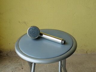 Handheld metal shower head lying on round stool against yellow wall background.