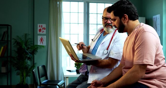 Indian male doctor using laptop while consulting young man patient as experienced physician discusses medical reports in modern Indian clinic OPD healthcare consultation
