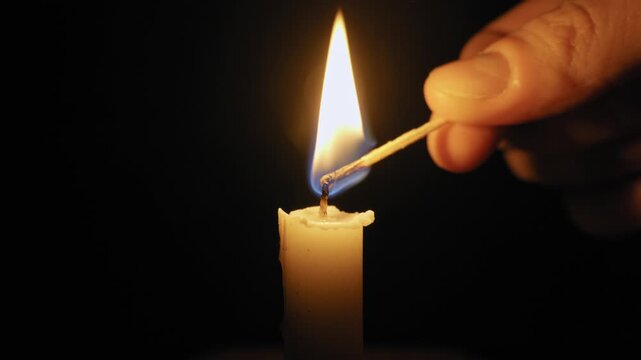 Hand with a lit match lights a single white candle against a black background. The warm flame is the only source of light, creating an intimate and peaceful mood.