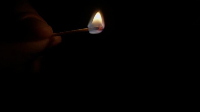 Hand strikes a wooden match against a matchbox, creating a small, bright flame. Extreme close-up shot with a completely black background highlights the ignition.