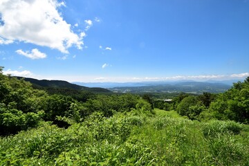A refreshing highland landscape view from Urabandai in Fukushima Prefecture, Japan, looking out over lush green grass and forests towards distant lakes and mountains under a bright blue sky.