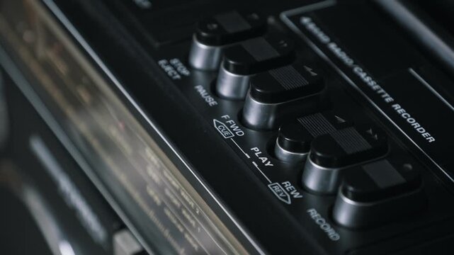 Close-up angled shot of a finger pressing the play button on a vintage cassette recorder. The dim lighting emphasizes the silver and black control keys of the analogue device.