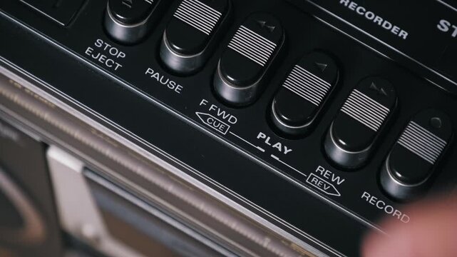Extreme close-up of a finger pressing play and stop keys on a vintage stereo player. The textured black buttons with chrome accents highlight the retro analog design.