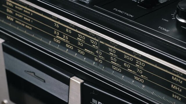 Wide macro shot of a radio tuner indicator scanning across an analog frequency scale. The detailed station markings for MW and SW bands highlight the vintage boombox design.