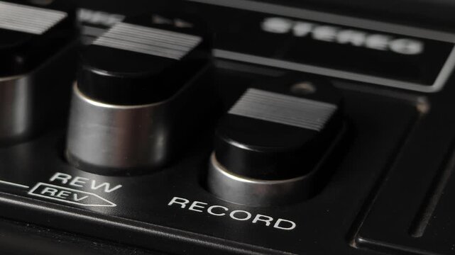 Macro shot of a finger pressing the record button on a vintage cassette deck. The textured black and silver key and adjacent rewind button highlight the analog audio interface.