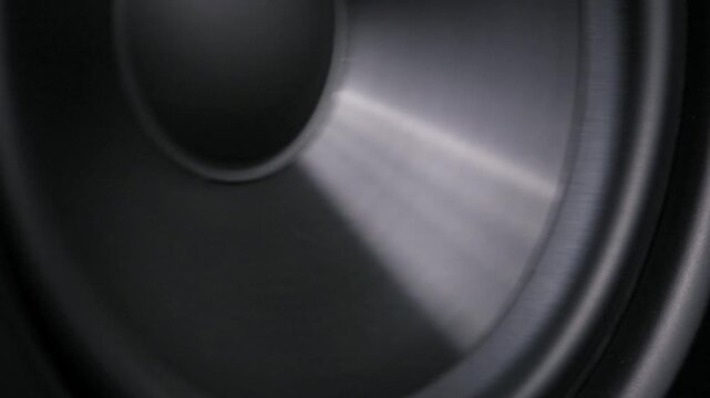 Extreme close-up macro view of a vibrating black subwoofer cone with a rubber surround. The speaker moves to deep bass frequencies during an acoustic studio test.