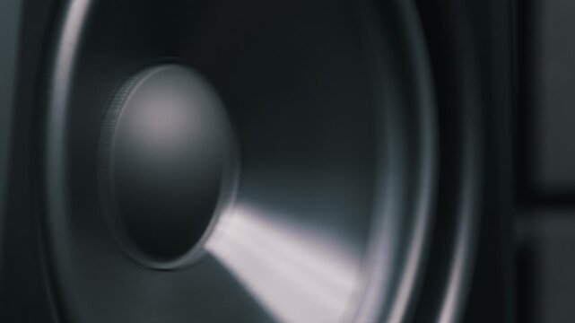 Close-up shot of a black speaker cone vibrating rhythmically to bass sounds. The subwoofer membrane moves back and forth, creating powerful sound waves in a dark studio.