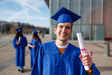 Male graduate student smiling, holding a diploma with classmates blurred in background