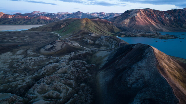 Aerial view of rugged peaks kissed by the fading light, their slopes cascading into serene waters, a tapestry of contrasting hues, Highlands, Iceland.