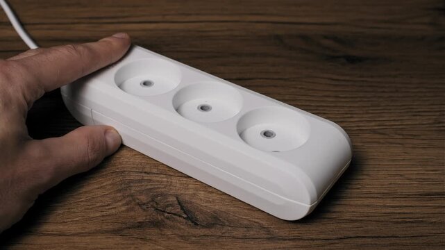 A hand unplugs a black two-pin adapter from a white triple socket extension cord on a dark wood table. Disconnecting the device from the electrical supply saves energy and money.