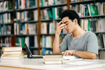 Focused stressed young man studying alone in a library using books and a tablet. The setting represents self improvement, lifelong learning, research work, and modern student lifestyle.