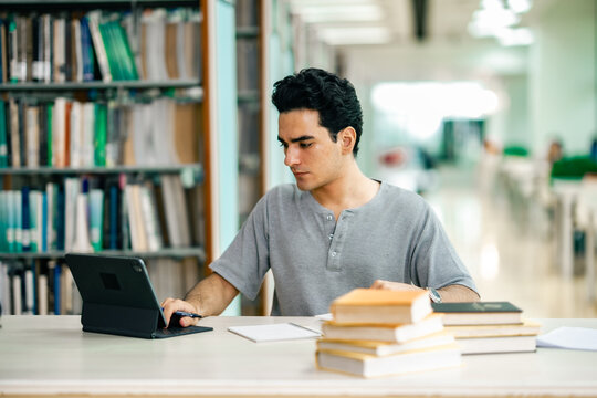 Male student studying in library with textbooks and tablet on table. The calm atmosphere, research preparation, higher education, and balance between traditional reading and digital learning. - Powered by Adobe