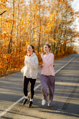 Fototapeta premium Two friends jog along a tree-lined road, surrounded by vibrant fall foliage. Their laughter fills the air as they share a fun moment on a bright, sunny day.