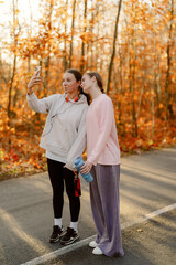 Fototapeta premium Two friends stand together on a winding road, surrounded by vibrant fall leaves, capturing joyful moments on their smartphones. They wear cozy attire and smile happily.