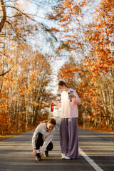 Fototapeta premium Two women stand on a quiet road covered in vibrant autumn leaves. One is tying her running shoes while the other holds a drink, both excited to start their workout in the crisp air.