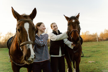 Fototapeta premium Two friends share smiles while standing by their horses in a serene field. The sun sets gently, casting warm light, creating a joyful atmosphere of friendship and nature.