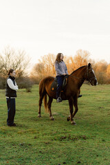 A girl sits confidently on a brown horse while an adult stands nearby, observing. The sun sets, casting a warm glow over the lush green field and surrounding trees.