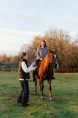 A woman rides a brown horse as another person guides her with gentle encouragement. The sun sets behind trees, creating a peaceful atmosphere for this outdoor riding lesson.