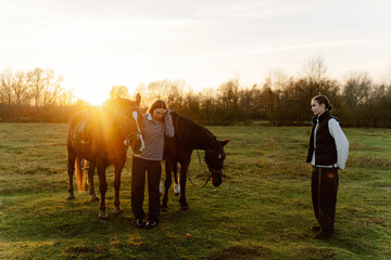 Two riders interact with their horses in a serene pasture as the sun sets, casting a warm glow. The peaceful scene captures a moment of bonding and connection in nature.