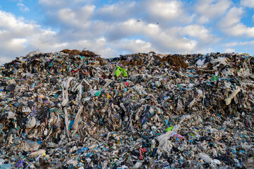 Garbage and waste cover a landfill site with plastic, debris, and other rubbish scattered widely under a cloudy sky during the day