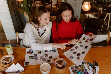 Two young women sit together at a wooden table in a cafe, excitedly discussing their travel ideas. They point at printed photos, sharing laughs and inspiration while enjoying drinks.