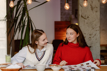 Two friends sit at a wooden table in a warm cafe, sharing laughter and ideas over bowls of snacks. They browse through a collection of design inspirations, surrounded by soft lighting.
