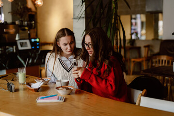 Obraz premium Two friends enjoy a lively conversation over drinks and dessert in a warm cafe. Laughter fills the air as they share stories and connect over their phones.