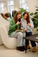 Obraz premium Two friends sit comfortably in a stylish café, flipping through a book and sharing animated conversations. Their laughter fills the air as they enjoy each other's company and explore new stories.