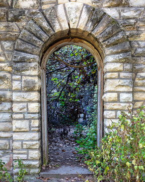 weathered stone archway leading into a narrow, overgrown garden path framed by rough-hewn stones and surrounded by lush greenery, creating a rustic and secluded atmosphere.