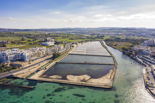Aerial view of shimmering turquoise waters embrace the rugged coastline near the distinctive salt pans, contrasting with the urban landscape, Bugibba, St. Paul's Bay, Malta.