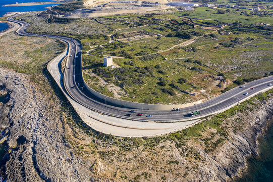 Aerial view of a winding coastal road slicing through rugged terrain where the turquoise sea meets the rocky coastline, Bugibba, St. Paul's Bay, Malta.