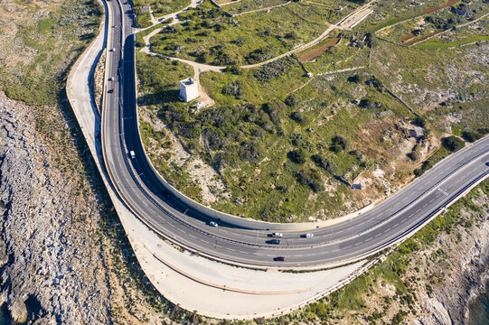 Aerial view of a winding highway hugging the rugged coastline, contrasting with the lush greenery inland, Bugibba, St. Paul's Bay, Malta.