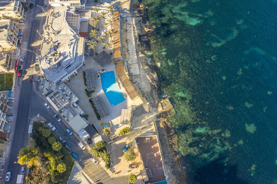 Aerial view of turquoise waters meet the rugged coastline near a hotel with a bright blue pool, Bugibba, St. Paul's Bay, Malta.