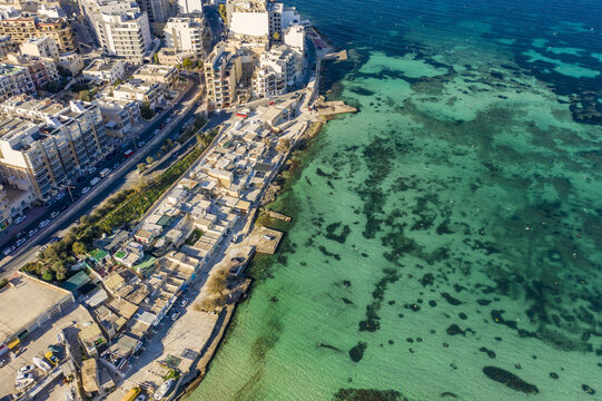 Aerial view of buildings meeting the turquoise sea, creating a striking contrast of urban development and natural beauty, Bugibba, St. Paul's Bay, Malta.
