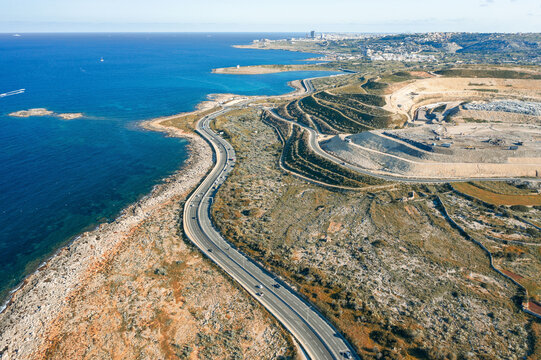 Aerial view of a winding coastal road contrasts sharply with the rugged cliffs and the azure sea near Bugibba, St. Paul's Bay, Malta.