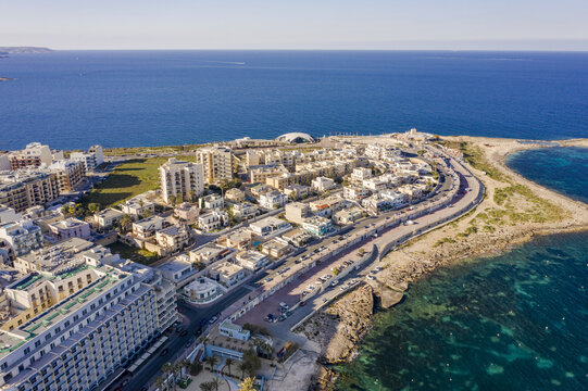 Aerial view of the bright, sun-drenched Bugibba coastline with rocky shores meeting the deep blue sea, buildings clustered along the coast, Bugibba, St. Paul's Bay, Malta.