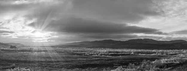 Dramatic northern landscape with sun rays breaking through clouds over wide tundra and distant mountains. Atmospheric black and white fine art scenery, calm, remote, and timeless.