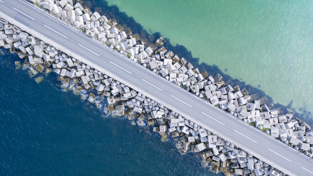 Aerial view of a road bisecting the turquoise and deep blue sea, flanked by geometric concrete blocks, creating a stark contrast of colors and textures, Kirkwall, Scotland, United Kingdom.