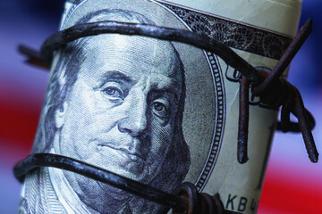 American dollar bill trapped in barbed wire against US flag, representing economic warfare, sanctions, financial pressure, and geopolitical conflict.