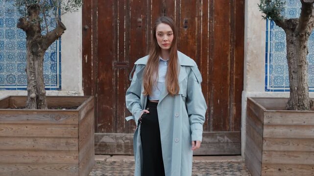 Fashionable young woman in blue coat adjusting long hair while standing near large wooden door in city during daytime