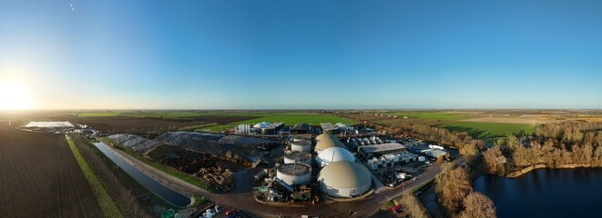 Aerial view of a sprawling industrial complex with rounded storage tanks, surrounded by fields and a reflective lake under a clear sky, Wisbech, England, United Kingdom.