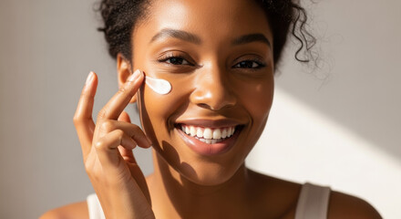 Obraz premium Close-up of a smiling young woman applying face cream with her finger. She has clear, radiant skin, in natural light against a neutral background.