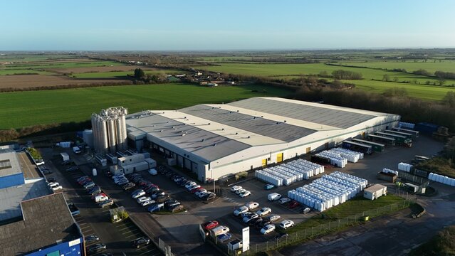 Aerial view of a large industrial building complex with rows of parked cars and stacked goods under a clear sky, Ely, England, United Kingdom.